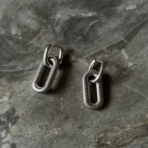 May include: A pair of silver-colored hoop earrings. Each earring features a textured, elongated oval design, connected to a small hoop closure. The earrings are displayed on a gray surface.