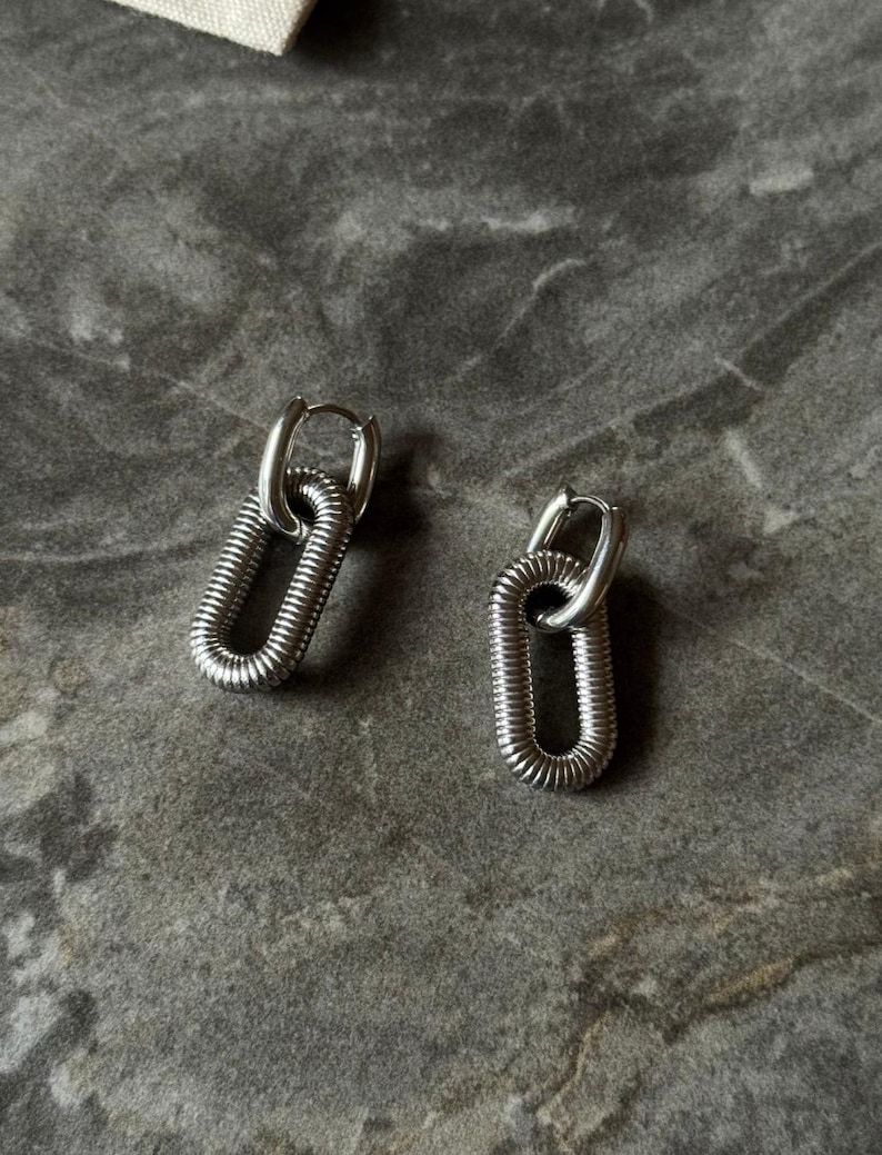 May include: A pair of silver-colored hoop earrings. Each earring features a textured, elongated oval design, connected to a small hoop closure. The earrings are displayed on a gray surface.