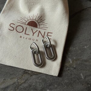 May include: A pair of silver-toned oval link earrings with a textured design. The earrings are displayed on a cream-colored pouch with the brand name "SOLYNE BIJOUX" printed in brown.