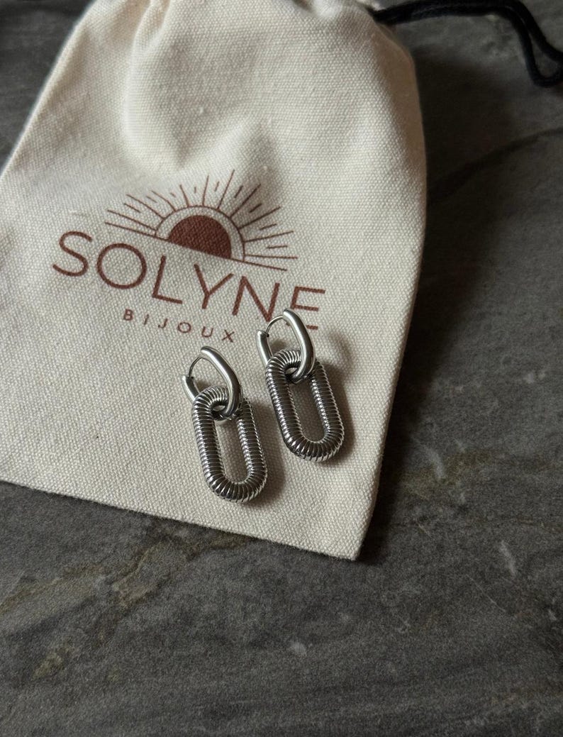 May include: A pair of silver-toned oval link earrings with a textured design. The earrings are displayed on a cream-colored pouch with the brand name "SOLYNE BIJOUX" printed in brown.
