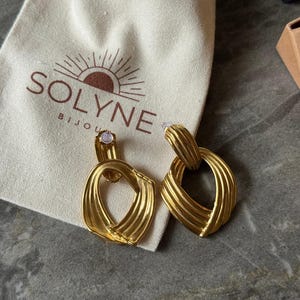May include: A pair of gold-colored earrings with a textured, geometric design. The earrings are displayed next to a cream-colored pouch with the brand name "SOLYNE BIJOUX" printed on it.