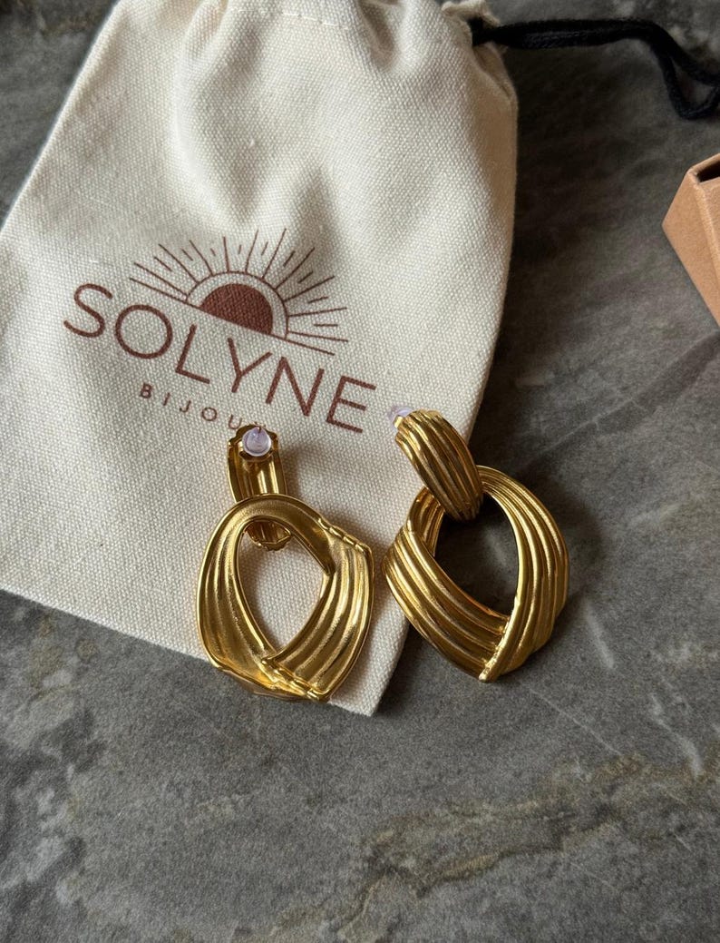 May include: A pair of gold-colored earrings with a textured, geometric design. The earrings are displayed next to a cream-colored pouch with the brand name "SOLYNE BIJOUX" printed on it.