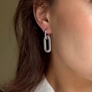 May include: Silver-toned chain link earrings. The earrings feature a textured, elongated oval design, hanging from a simple hoop. The jewelry is shown against a neutral background, highlighting the metallic shine.