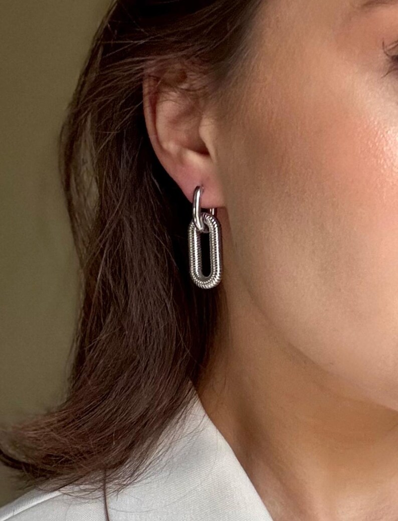May include: Silver-toned chain link earrings. The earrings feature a textured, elongated oval design, hanging from a simple hoop. The jewelry is shown against a neutral background, highlighting the metallic shine.