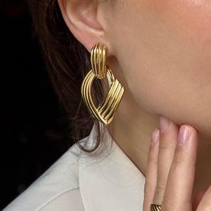May include: Gold-toned earrings with a geometric design. The earrings feature a combination of curved and linear elements, creating a layered effect. The design includes a rounded top section and a diamond-shaped bottom section. A matching gold ring is also visible.