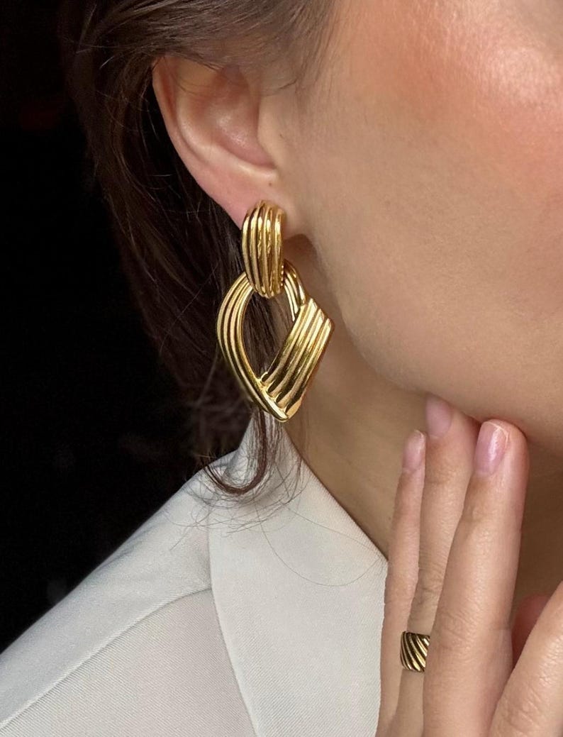 May include: Gold-toned earrings with a geometric design. The earrings feature a combination of curved and linear elements, creating a layered effect. The design includes a rounded top section and a diamond-shaped bottom section. A matching gold ring is also visible.