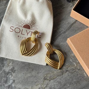 May include: A pair of gold-colored earrings with a woven design, resting on a cream-colored pouch with the brand name "SOLYME BIJOUX" printed on it. A small, open cardboard box is in the background. The earrings have a textured, geometric shape.