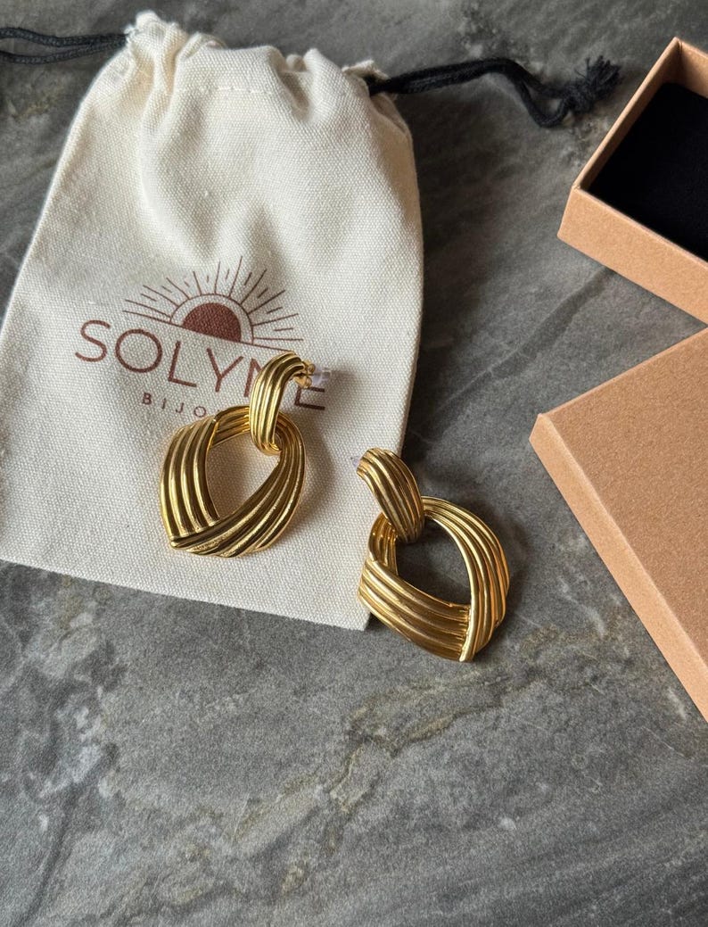 May include: A pair of gold-colored earrings with a woven design, resting on a cream-colored pouch with the brand name "SOLYME BIJOUX" printed on it. A small, open cardboard box is in the background. The earrings have a textured, geometric shape.