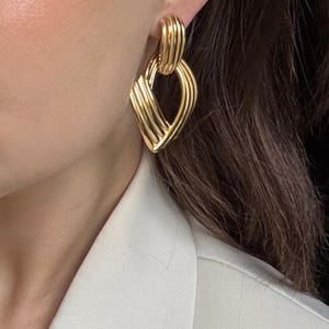 May include: Gold-toned earrings with a geometric design. The earrings feature a ribbed texture and a diamond-shaped pendant. The earrings are worn by a person wearing a white blazer.