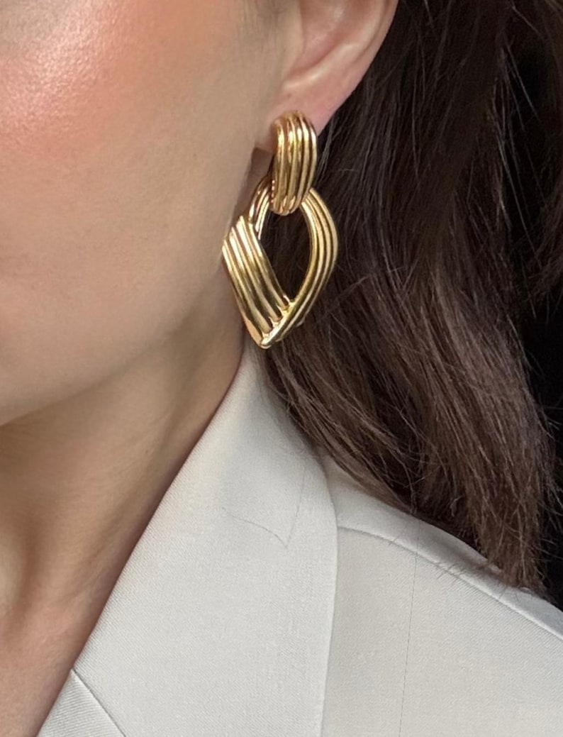 May include: Gold-toned earrings with a geometric design. The earrings feature a ribbed texture and a diamond-shaped pendant. The earrings are worn by a person wearing a white blazer.