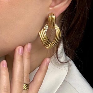 May include: Gold-toned earrings with a geometric design. The earrings feature a rectangular top and a diamond-shaped bottom, both with a ribbed texture. A matching gold ring is also visible.