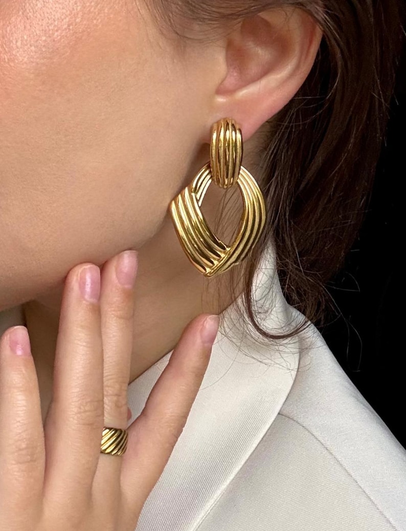 May include: Gold-toned earrings with a geometric design. The earrings feature a rectangular top and a diamond-shaped bottom, both with a ribbed texture. A matching gold ring is also visible.