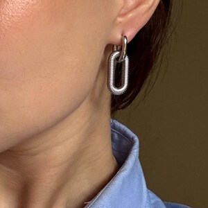 May include: Silver-toned chain link earrings. The earrings feature a textured, elongated oval design, with a smaller loop connecting to the ear. The earrings are shown against a light blue collared shirt.
