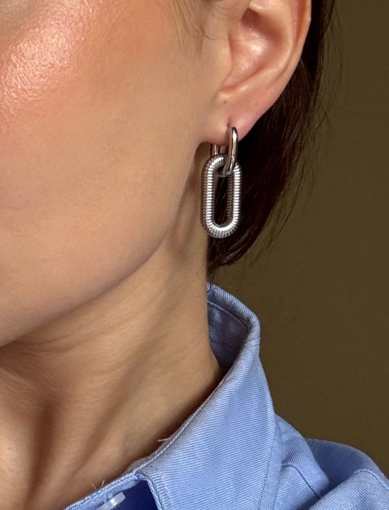 May include: Silver-toned chain link earrings. The earrings feature a textured, elongated oval design, with a smaller loop connecting to the ear. The earrings are shown against a light blue collared shirt.