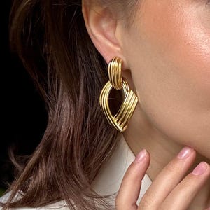 May include: Gold-toned earrings with a geometric design. The earrings feature a series of parallel lines and curves, creating a layered, textured appearance. The earrings are worn on an ear, and a matching ring is visible on a finger.