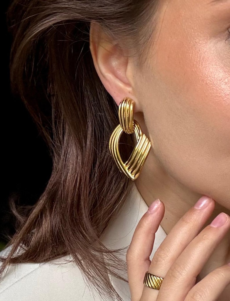 May include: Gold-toned earrings with a geometric design. The earrings feature a series of parallel lines and curves, creating a layered, textured appearance. The earrings are worn on an ear, and a matching ring is visible on a finger.