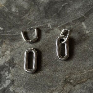 May include: Silver-toned hoop earrings and two oval-shaped earrings. The earrings have a textured, ribbed design. The earrings are displayed on a gray, textured surface.
