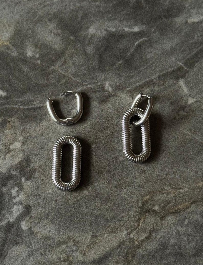 May include: Silver-toned hoop earrings and two oval-shaped earrings. The earrings have a textured, ribbed design. The earrings are displayed on a gray, textured surface.