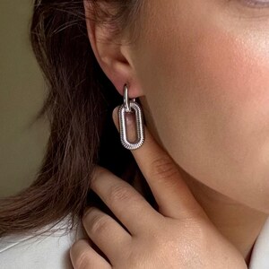 May include: Silver-toned, rectangular link earrings. The earrings are connected by a small hoop that goes through the ear. The rectangular links have a textured, ridged design. The earrings are worn on a person.