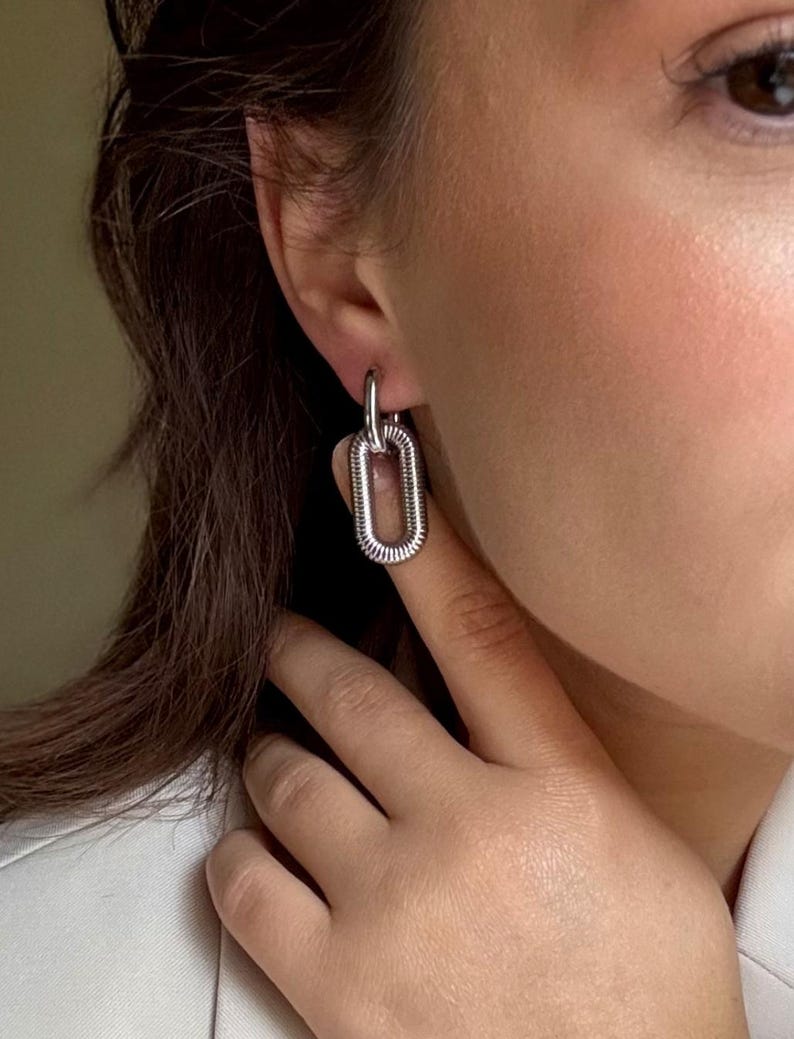 May include: Silver-toned, rectangular link earrings. The earrings are connected by a small hoop that goes through the ear. The rectangular links have a textured, ridged design. The earrings are worn on a person.