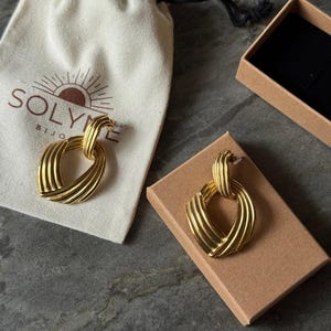 May include: Gold-toned earrings with a textured, woven design, displayed on a brown cardboard box and a cream-colored drawstring bag. The bag has the brand name "SOLYME BIJOU" printed on it. The earrings are a statement piece.