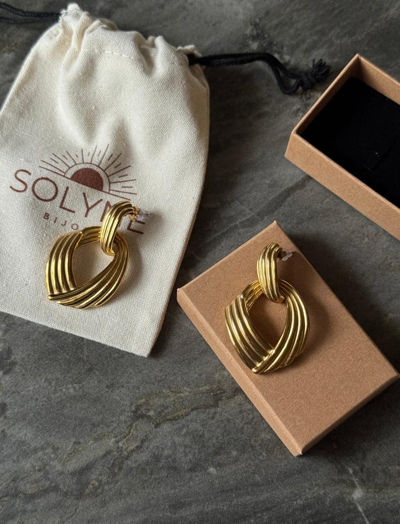 May include: Gold-toned earrings with a textured, woven design, displayed on a brown cardboard box and a cream-colored drawstring bag. The bag has the brand name "SOLYME BIJOU" printed on it. The earrings are a statement piece.