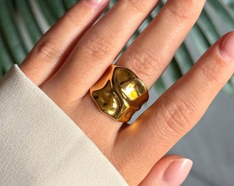 May include: A gold-colored ring with an irregular, textured surface. The ring is worn on a hand with manicured nails and a white sleeve in the background. The ring's design is abstract and sculptural, reflecting light.