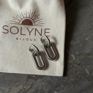 May include: A pair of silver-colored hoop earrings with an oval, coiled design. The earrings are displayed on a cream-colored pouch with the brand name "SOLYNE BIJOUX" printed in brown. The background is a gray, textured surface.