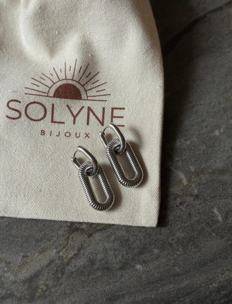 May include: A pair of silver-colored hoop earrings with an oval, coiled design. The earrings are displayed on a cream-colored pouch with the brand name "SOLYNE BIJOUX" printed in brown. The background is a gray, textured surface.