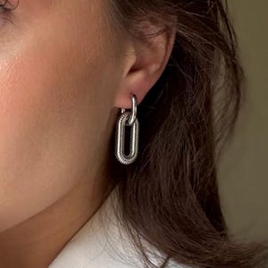 May include: Silver-toned chain link earrings. The earrings feature a textured, oval-shaped design, with a smaller loop connecting to the ear. The jewelry is shown on a person's ear, with a neutral background.