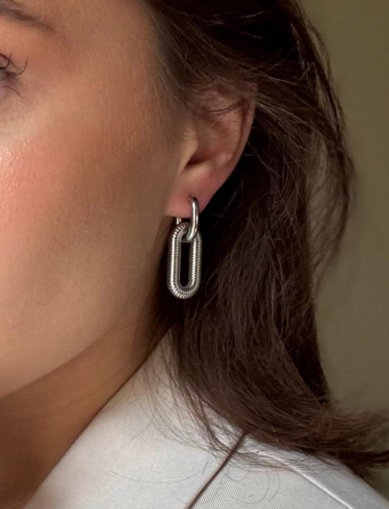 May include: Silver-toned chain link earrings. The earrings feature a textured, oval-shaped design, with a smaller loop connecting to the ear. The jewelry is shown on a person's ear, with a neutral background.