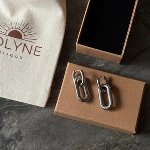May include: A pair of silver-colored chain link earrings on a brown cardboard box. The earrings are next to a small beige bag with the text "OLYNE BIJOUX" printed on it. An open brown box with a black interior is also in the image.