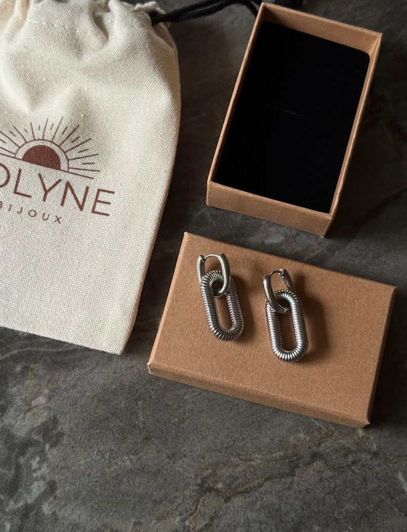 May include: A pair of silver-colored chain link earrings on a brown cardboard box. The earrings are next to a small beige bag with the text "OLYNE BIJOUX" printed on it. An open brown box with a black interior is also in the image.