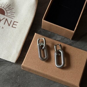 May include: A pair of silver-colored chain link earrings with a textured design. The earrings are displayed on a brown cardboard box next to a small canvas bag with the word "YNE" printed on it.