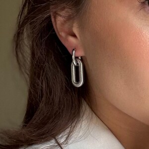 May include: Silver-toned chain link hoop earrings. The earrings feature a textured, elongated oval design, with a smaller hoop connecting to the ear. The jewelry is worn on a person's ear, with a white blazer in the background.