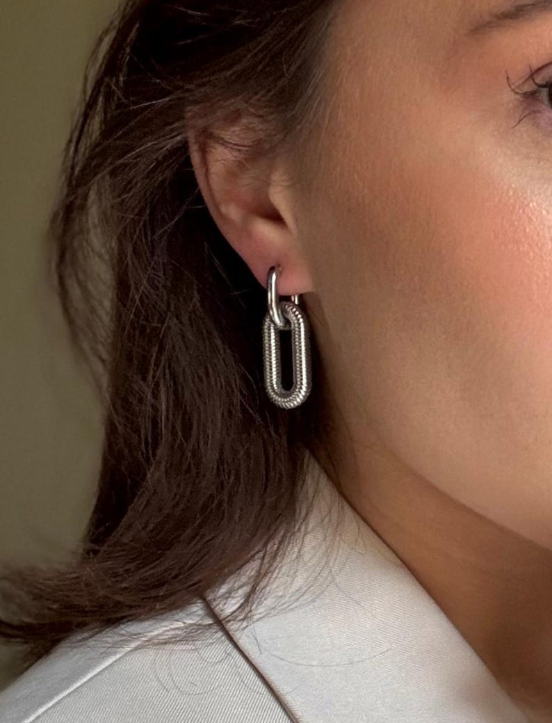 May include: Silver-toned chain link hoop earrings. The earrings feature a textured, elongated oval design, with a smaller hoop connecting to the ear. The jewelry is worn on a person's ear, with a white blazer in the background.