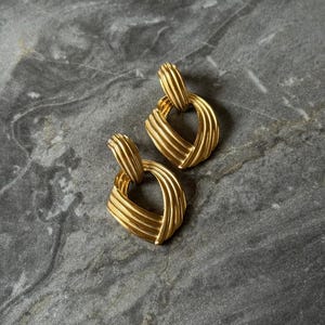May include: A pair of gold-toned, square-shaped earrings. Each earring features a textured, ridged design, with a loop at the top for attachment. The earrings are displayed on a gray, marbled surface.