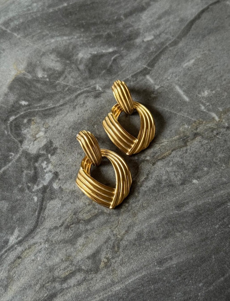 May include: A pair of gold-toned, square-shaped earrings. Each earring features a textured, ridged design, with a loop at the top for attachment. The earrings are displayed on a gray, marbled surface.