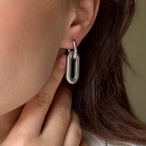 May include: Silver-toned chain link earrings. The earrings are a polished silver color and have a textured, ribbed design. The earrings are a hoop style and are being worn.
