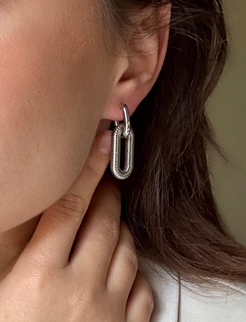 May include: Silver-toned chain link earrings. The earrings are a polished silver color and have a textured, ribbed design. The earrings are a hoop style and are being worn.