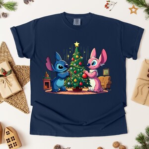 May include: Navy blue t-shirt featuring a cartoon illustration of two characters decorating a Christmas tree. One character is blue, the other is pink. The tree is adorned with ornaments and a star. The shirt is surrounded by holiday decorations.