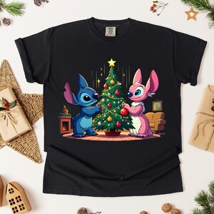 May include: Black t-shirt featuring a cartoon illustration of two characters decorating a Christmas tree. The characters are blue and pink, with the tree adorned with ornaments. The design is set against a backdrop of a room with a fireplace and a couch.