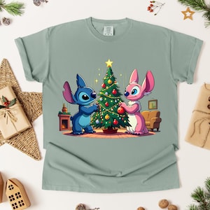 May include: A light green t-shirt featuring a cartoon illustration of two characters decorating a Christmas tree. One character is blue, the other is pink. The tree is adorned with ornaments and lights. The shirt is surrounded by Christmas decorations.