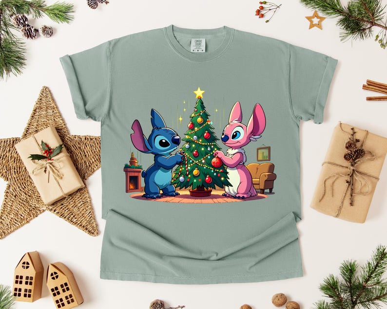 May include: A light green t-shirt featuring a cartoon illustration of two characters decorating a Christmas tree. One character is blue, the other is pink. The tree is adorned with ornaments and lights. The shirt is surrounded by Christmas decorations.
