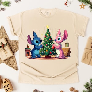 May include: Beige t-shirt featuring a cartoon illustration of Stitch and Angel decorating a Christmas tree. The characters are blue and pink, with the tree adorned with ornaments. The shirt is surrounded by Christmas decorations.