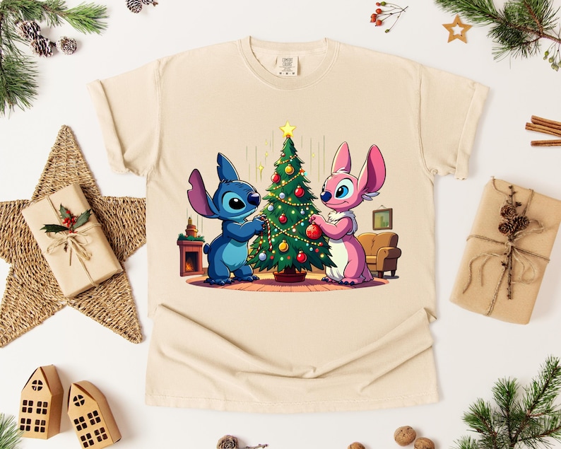 May include: Beige t-shirt featuring a cartoon illustration of Stitch and Angel decorating a Christmas tree. The characters are blue and pink, with the tree adorned with ornaments. The shirt is surrounded by Christmas decorations.