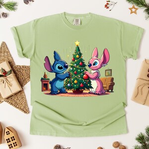 May include: Light green t-shirt featuring a cartoon illustration of two characters decorating a Christmas tree. One character is blue, the other is pink. The tree is decorated with ornaments and a star. The shirt is surrounded by Christmas decorations.