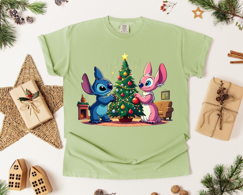 May include: Light green t-shirt featuring a cartoon illustration of two characters decorating a Christmas tree. One character is blue, the other is pink. The tree is decorated with ornaments and a star. The shirt is surrounded by Christmas decorations.