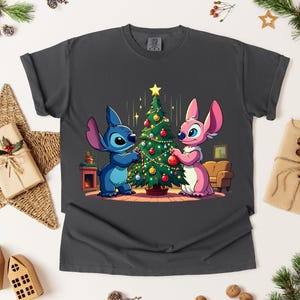 May include: Dark gray t-shirt featuring a cartoon illustration of two characters decorating a Christmas tree. One character is blue, the other is pink. The tree is adorned with ornaments and lights, set against a cozy interior scene.