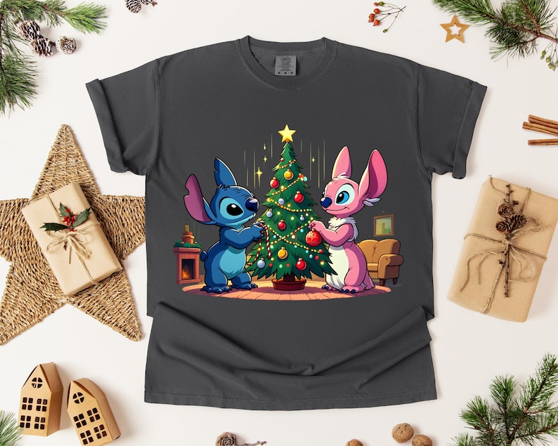 May include: Dark gray t-shirt featuring a cartoon illustration of two characters decorating a Christmas tree. One character is blue, the other is pink. The tree is adorned with ornaments and lights, set against a cozy interior scene.